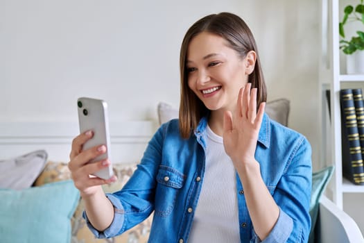 Young woman sitting with smartphone on couch at home having video call. Smiling female chatting with friends colleagues on social networks, student talking in online lesson freelancer working remotely