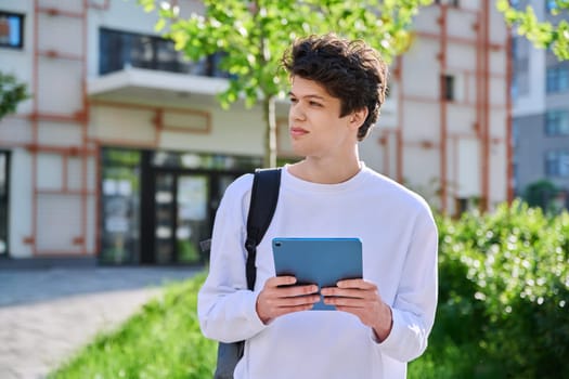 Young handsome guy college student using digital tablet outdoor, educational building background. Education, technology, training, 19,20 years age youth concept