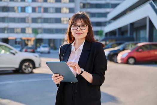 Mature confident successful business woman with digital tablet in black suit looking at camera outdoors, backdrop of modern city. Business, entrepreneurship mentoring insurance sales advertising work