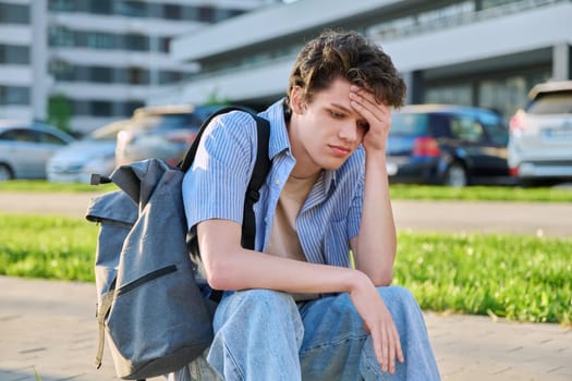 Upset sad unhappy young male sitting outdoor on steps. Guy university college student with backpack sitting, holding head with hands. Problems, difficulties, depression, mental health of young people