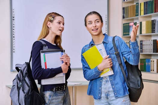 Teenage female students talking in a library class. Smiling teenagers from their classmates with textbooks and backpacks. High school education adolescence friendship communication concept