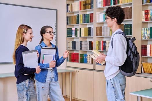 Group of teenage high school students in library classroom. Three schoolchildren 16, 17 years old, talking laughing together. Education, adolescence, friendship communication concept