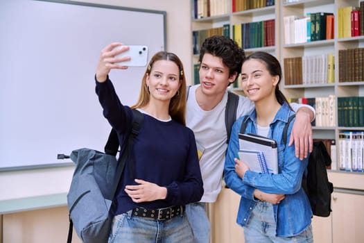 Selfie portrait of group friends teenagers high school students looking at camera inside classroom. Having fun cheerful teenage guy and two girls in library. Friendship adolescence education lifestyle