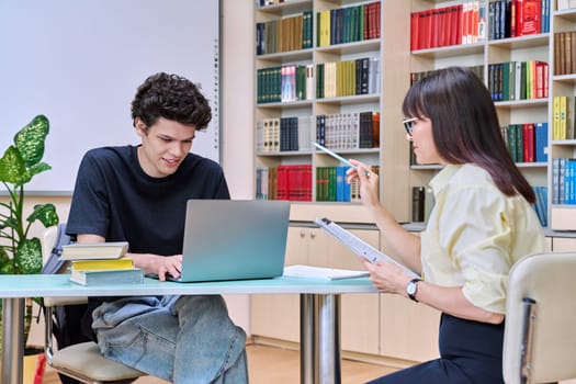 Young male college student talking studying with female teacher. Mentor and guy together in educational library office room. Education, training, mentoring, teaching, youth concept