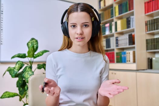 Webcam view of teenage girl, high school student, in headphones, talking to camera. Female teenager studying remotely, video conference chat, online test exam lesson. Technology education adolescence