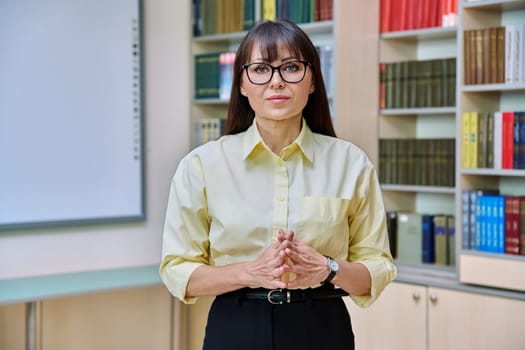 Portrait of confident mature business woman in library. Middle-aged elegant female with folded hands, manager teacher mentor psychologist counselor librarian advisor background of shelves with books