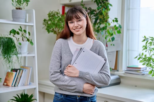 Portrait of smiling female student, 19,20 years old, looking at camera holding notebooks and textbooks, in home interior. Youth, lifestyle, education college university concept