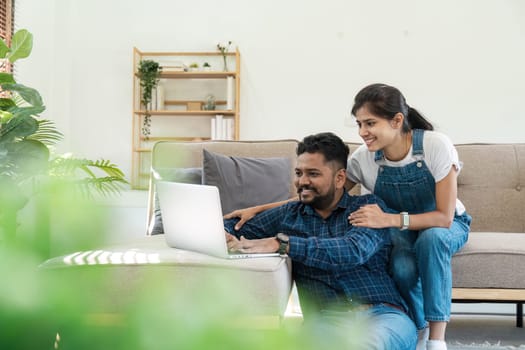 Indian couple sitting in a modern living room, using a laptop to pay bills and plan their finances together.