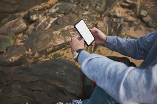 A man holding a smartphone with a blank screen, sitting on rocky ground outdoors. He is wearing a gray hoodie and jeans, enjoying a casual moment in nature.