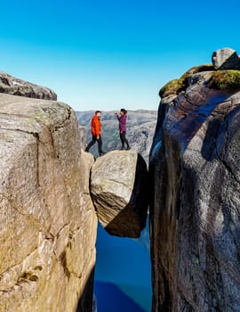 Two people stand on a rock bridge overlooking a stunning Norwegian landscape, seemingly suspended in mid-air. a couple of men and women visiting Kjeragbolten, Norway