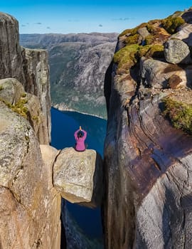 A person sits on the edge of Kjeragbolten, Norway, a famous cliff in Norway, with a view of the surrounding landscape and the fjord below. Asian woman at Kjeragbolten, Norway
