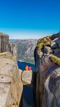 Two individuals sit on a narrow rock ledge overlooking a deep valley in Norway. The majestic Kjeragbolten cliff provides a breathtaking backdrop for this daring feat. a couple at Kjeragbolten