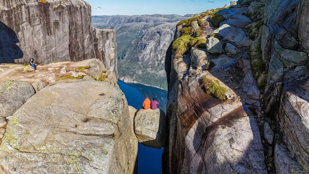 Two hikers sit on the edge of Kjeragbolten Pulpit Rock in Norway, enjoying the stunning panoramic view of the surrounding mountains and fjord. a couple of men and women at Kjeragbolten