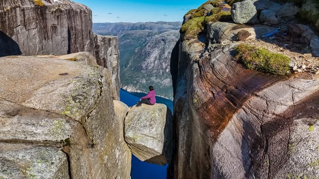 A lone hiker sits on the edge of Kjeragbolten, Norway, a famous cliff in Norway, overlooking a breathtaking fjord.