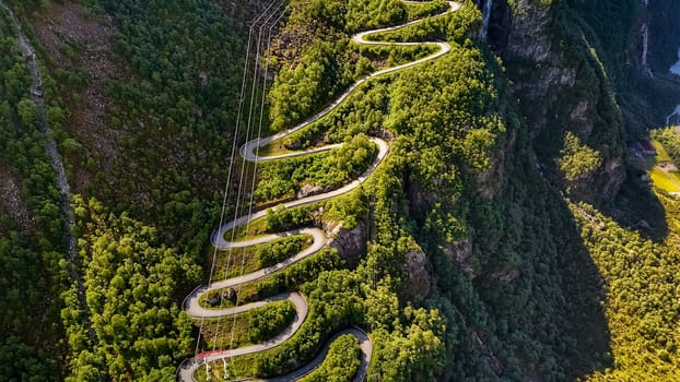 An aerial view of a winding road through the mountains of Norway. The road is surrounded by lush green trees and slopes down into a valley below. Lysebotn, Lysefjord, Norway