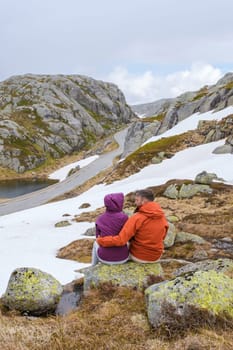 A couple sits on a rock, enjoying the breathtaking view of a winding road through snow-capped mountains in Norway.