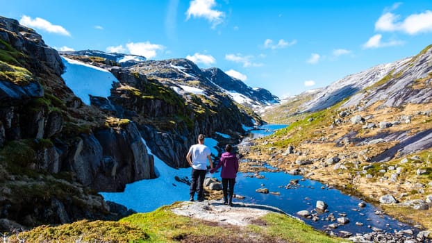Two hikers stand hand-in-hand on a rocky outcropping overlooking a pristine mountain lake in Norway, surrounded by snow-capped peaks and lush greenery. Kjeragbolten Norway