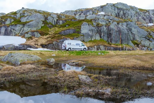 A white camper van is parked on a mountain road in Norway, with a reflection of the van in a small pond in the foreground.