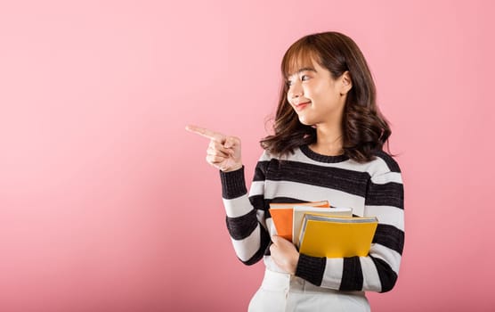 Portrait of happy Asian beautiful young woman confident smiling holding orange book open pointing finger to side copy space, studio shot isolated on pink background, education concept
