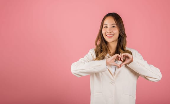 A confident business woman wear suit smiles and creates a heart symbol with two hands on white background. Asian portrait of a beautiful young female expressing love and happiness for Valentine's Day