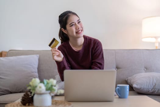 A young woman sitting on a sofa, holding a credit card and using a laptop for online shopping in a cozy home environment.