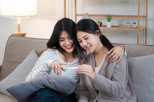 Two happy gay lesbian lgbt women couple sitting on sofa celebrating positive pregnancy test. lgbtq concept .