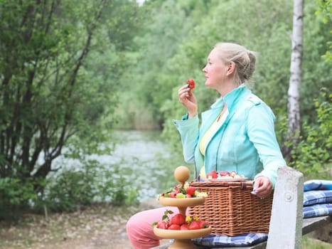 Portrait of a beautiful middle-aged caucasian blonde woman eating a big red and ripe strawberry sitting on a bench with a wicker basket in the forest alone resting from family and household chores,close-up side view. Concept picnic, outdoor recreation, relaxing mom, moms, happy woman, fruit barbecue, healthy food.