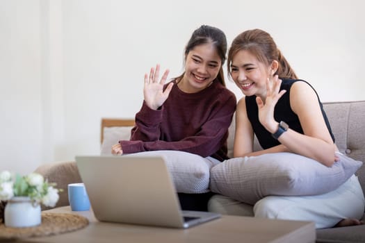 A joyful lesbian couple sitting on a couch, waving at a laptop screen during a video call. LGBTQ representation, modern technology, and love.