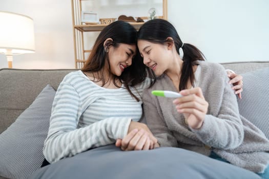 Two happy gay lesbian lgbt women couple sitting on sofa celebrating positive pregnancy test. lgbtq concept .