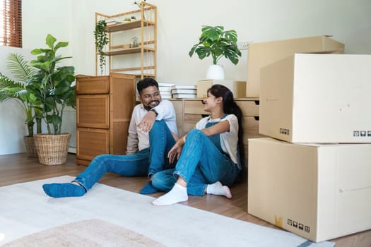 Indian couple moving into a new home, unpacking boxes, and relaxing in a modern living room with green plants and wooden furniture.