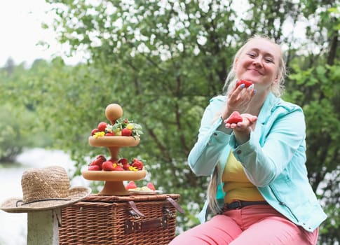 Portrait of a beautiful Caucasian cheerful middle-aged blonde woman eating a large ripe strawberry with appetizing pleasure, holding it out in her hand directly to the camera, sitting in a park on a bench on a spring day, close-up side view. Outdoor picnic concept, healthy eating and lifestyle.