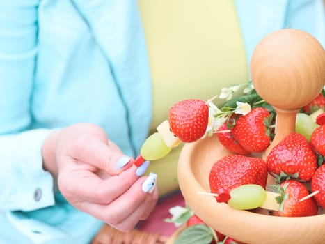 The hands of a Caucasian young unrecognizable woman with a beautiful delicate manicure in a blue jacket and a yellow T-shirt hold a fruit barbecue on a skewer of strawberry, cheese and wongrad next to a two-tiered wooden plate, close-up side view. Conceptual picnic, healthy eating.