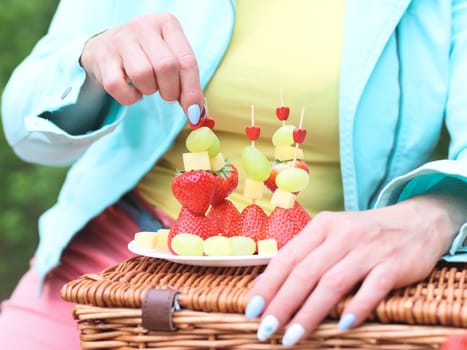 One Caucasian young unrecognizable blonde woman with delicate manicure in a blue jacket and yellow T-shirt puts ready-made fruit barbecue on a skewer of strawberries, cheese and wongrad on a plate, sitting in the park on a spring day, side view close-up. Spring picnic concept, healthy eating.