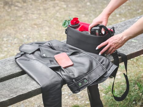 The hands of a young Caucasian woman with a beautiful delicate manicure take out a camera with a lens and a red rose from her purse on a wooden bench with a black leather jacket and a pink smartphone in a public park, close-up side view. Concept using technology, photography, theft.