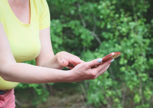 One young Caucasian unrecognizable woman holds a mobile phone in her hands, poking her finger at it while standing in the park on a sunny summer day, close-up side view. Concept of using technology.