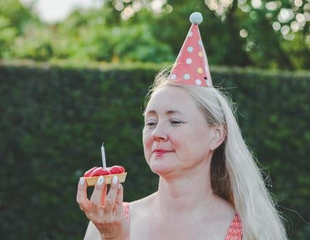 Portrait of a beautiful blonde middle-aged Caucasian woman with a cone on her head, a strawberry tartlet with a candle in her hands, celebrating her birthday with a smile, sitting on a wooden bench in the backyard of her house, close-up side view. Birthday celebration concept.