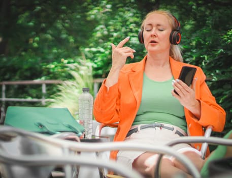 A young beautiful stylish Caucasian blonde woman in a bright orange jacket wearing headphones with a smartphone in her hands, meditating, listening to music, sitting with her legs stretched out on a chair in a street cafe on a summer day, close-up side view. Technology use concept.