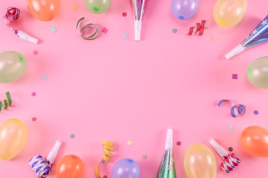 Many colored pastel inflated mini balloons with scattered confetti, tinsel and whistles lie in an oval frame on a pink background with copy space in the center, flat lay close-up.