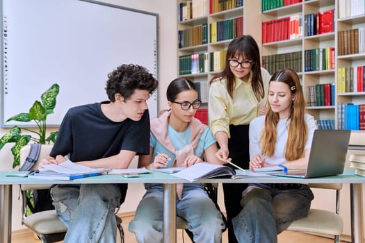 Group of teenage students sitting at desk with female mentor teacher study together prepare for tests inside classroom library office. High school, education, teamwork, learning, adolescence concept