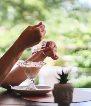 Hands of a young Caucasian unrecognizable woman eating chocolate ice cream with a wafer from a beautiful glass while sitting at a table in a street cafe, close-up side view. Concept of summer food, restaurant ice cream.