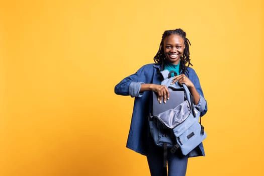 Portrait of cheerful woman putting laptop in backpack, preparing to go to school exam, isolated over studio background. Joyful pupil packing notebook in rucksack, preparing for university assessment