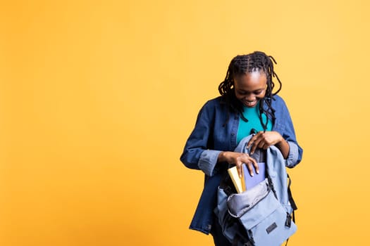 Joyful african american woman browsing through backpack, isolated over yellow studio background. Jolly student taking out school textbooks out of rucksack used during commuting to university