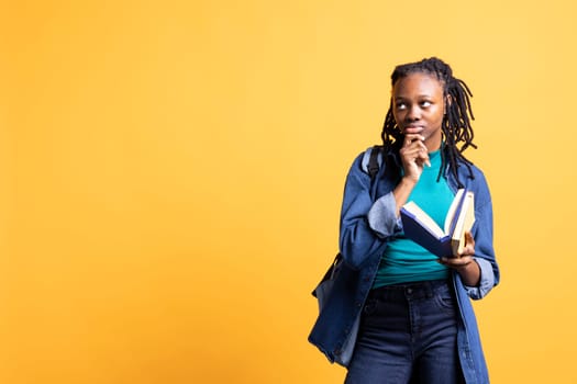 African american woman with book in hands deep in thoughts, isolated over yellow studio background. Bookworm holding novel, thinking about themes and characters after reading