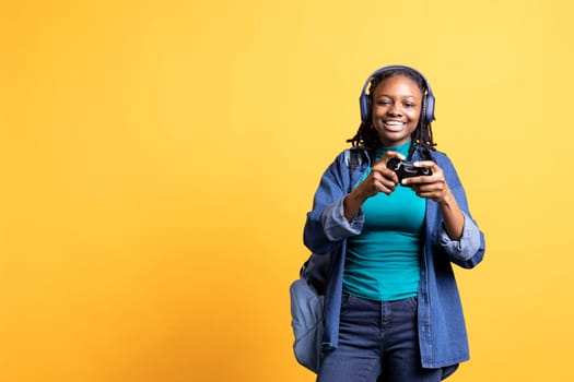 Portrait of happy teenager playing videogames with controller and headphones, studio background. Joyous gamer having fun participating in PvP internet game with joypad, relaxing after school