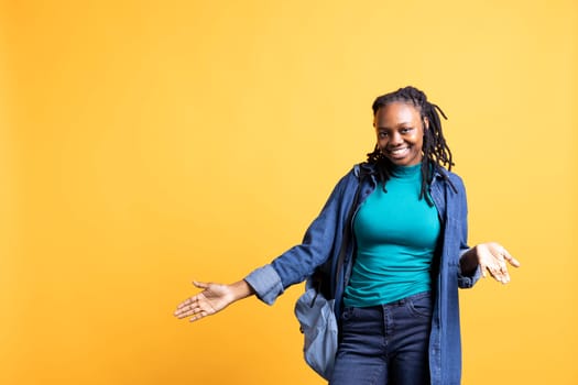 Happy young woman doing shrugging shoulders gesture, showing relaxed mood. Serene BIPOC girl doing nonchalant carefree hand gesturing, isolated over yellow studio background