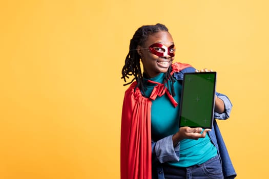 Portrait of smiling person portraying enigmatic superhero wearing mask, showcasing green screen tablet, studio background. Cheerful girl posing as hero with secret identity showing chroma key device