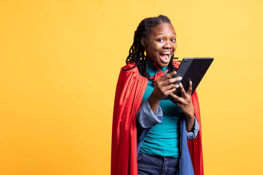 Portrait of ecstatic african american girl portraying superhero excitedly using tablet, studio background. Delighted woman posing as hero in costume, using digital device, having fun