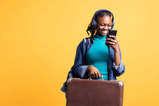 Upbeat tourist going on holiday with suitcase, phone and headphones. Traveler using cellphone to chat with mates, listening music, doing tourism, isolated over studio background