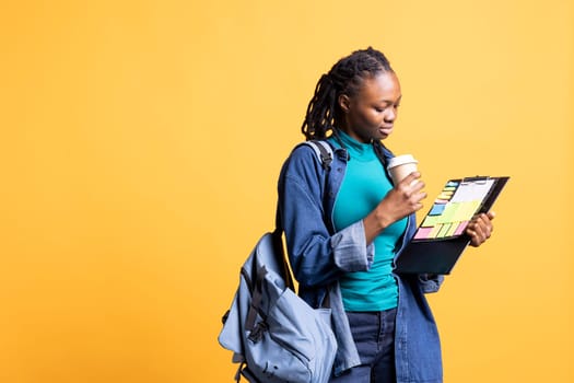 Woman with backpack sipping coffee while examining clipboard with colorful sticky notes against bright yellow backdrop. Organized person doing productivity tasks holding beverage cup