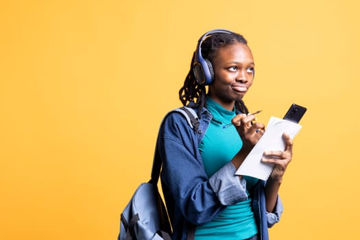 BIPOC student thinking how to solve homework exercises while listening music, isolated over studio background. Girl studying for exam, contemplating and enjoying songs on headphones